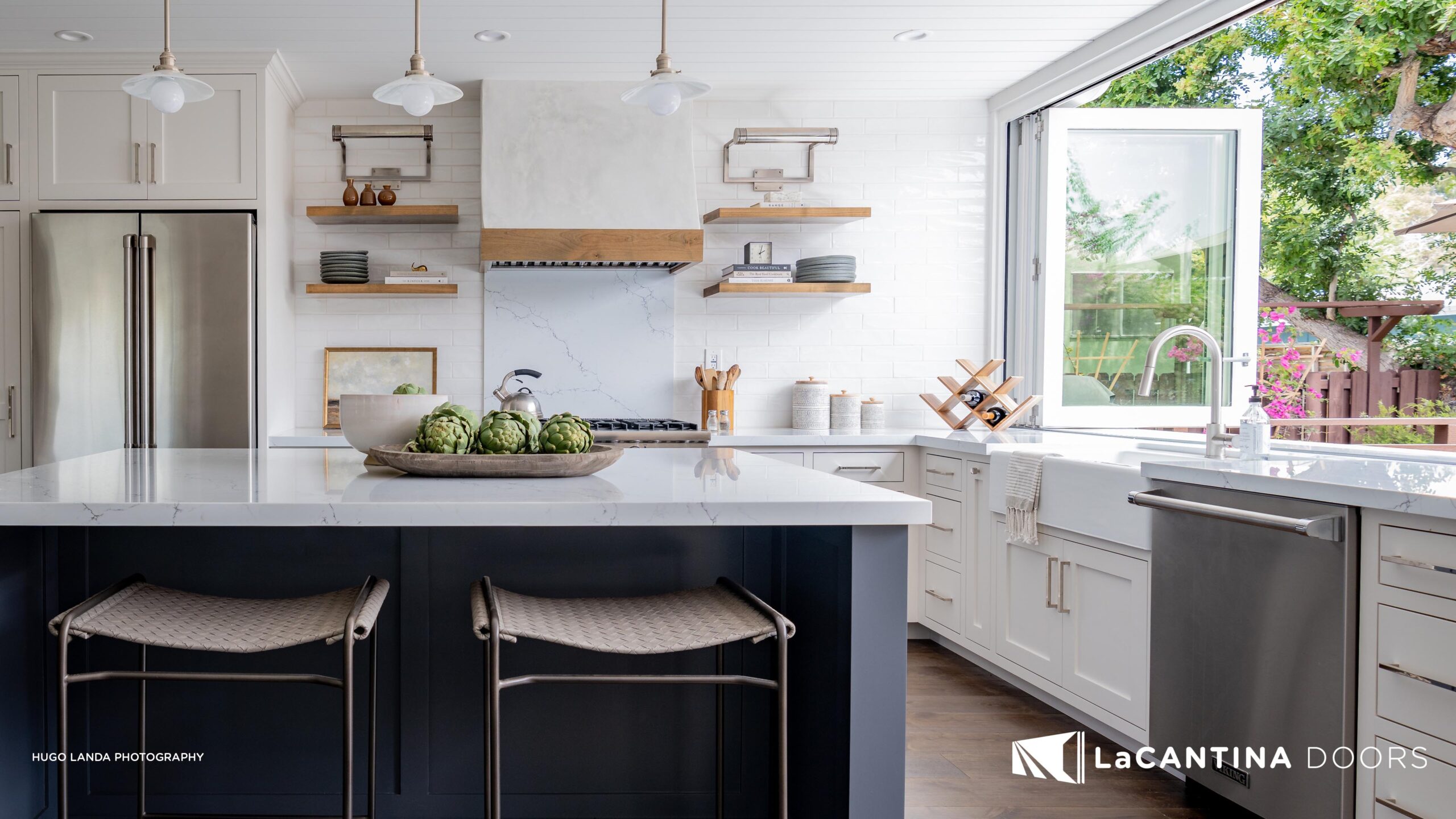 Modern kitchen with white cabinetry, an island with a dark blue base and gray bar stools, stainless steel appliances, and a large open window showcasing a garden view. LaCantina Doors branding visible.