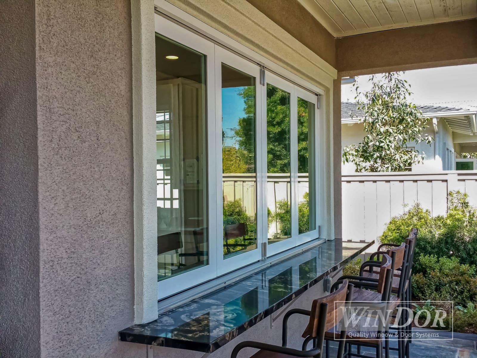 A large sliding window installed on the exterior wall of a house, with three barstools placed in front. The window frame is white, and there is a logo in the bottom right corner that reads "WIN-DOR.